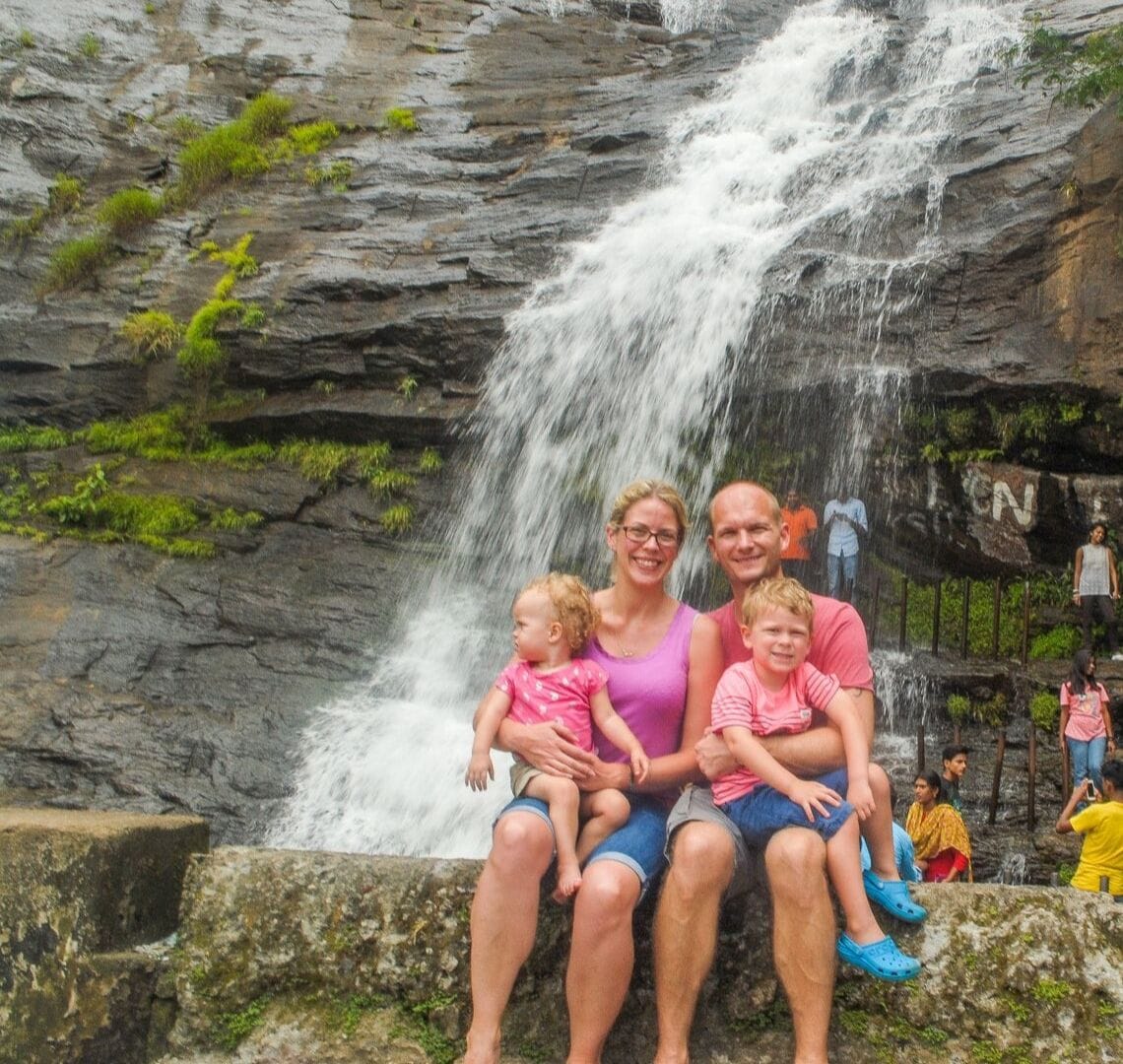 Wanderlust family photo in front of a waterfall on the way to Munnar. There are people in the background and Thing 2 is looking over her shoulder while the rest of us are looking at the camera. 