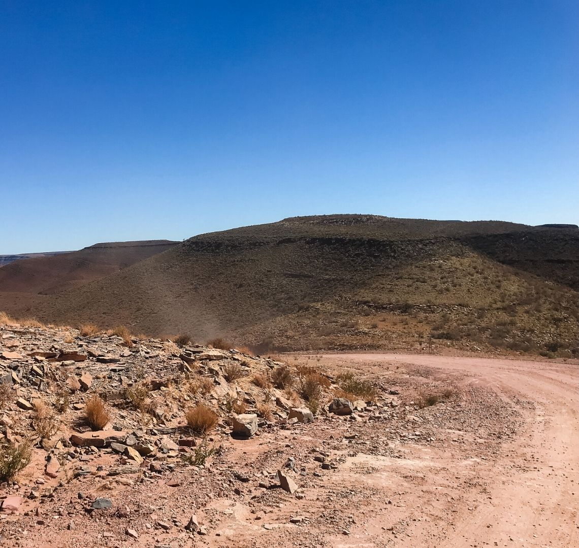 5 Things You Need To Know Before A Self-Drive Safari In Namibia - dust is kicked up behind a (hidden) car in front of us on a red dusty gravel track. The sky is completely blue. 