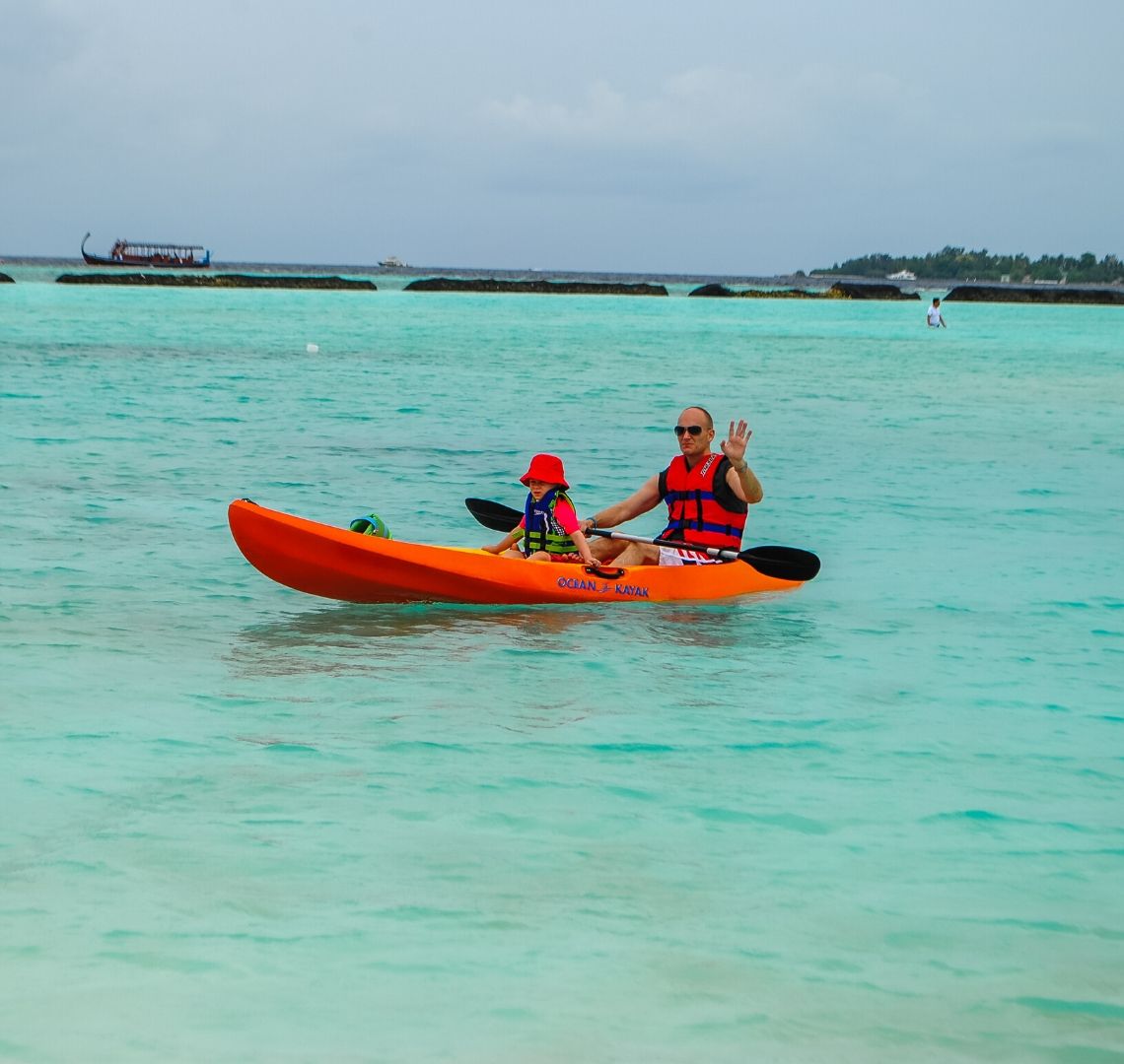Maldives Activities / Things to do in the Maldives - Thing 1 and Mr Wanderlust in bright orange canoe. Mr wanderlust is waving at me. The sea is bright blue and there is a traditional boat in the background behind the breakwater. The sky is cloudy and stormy. 