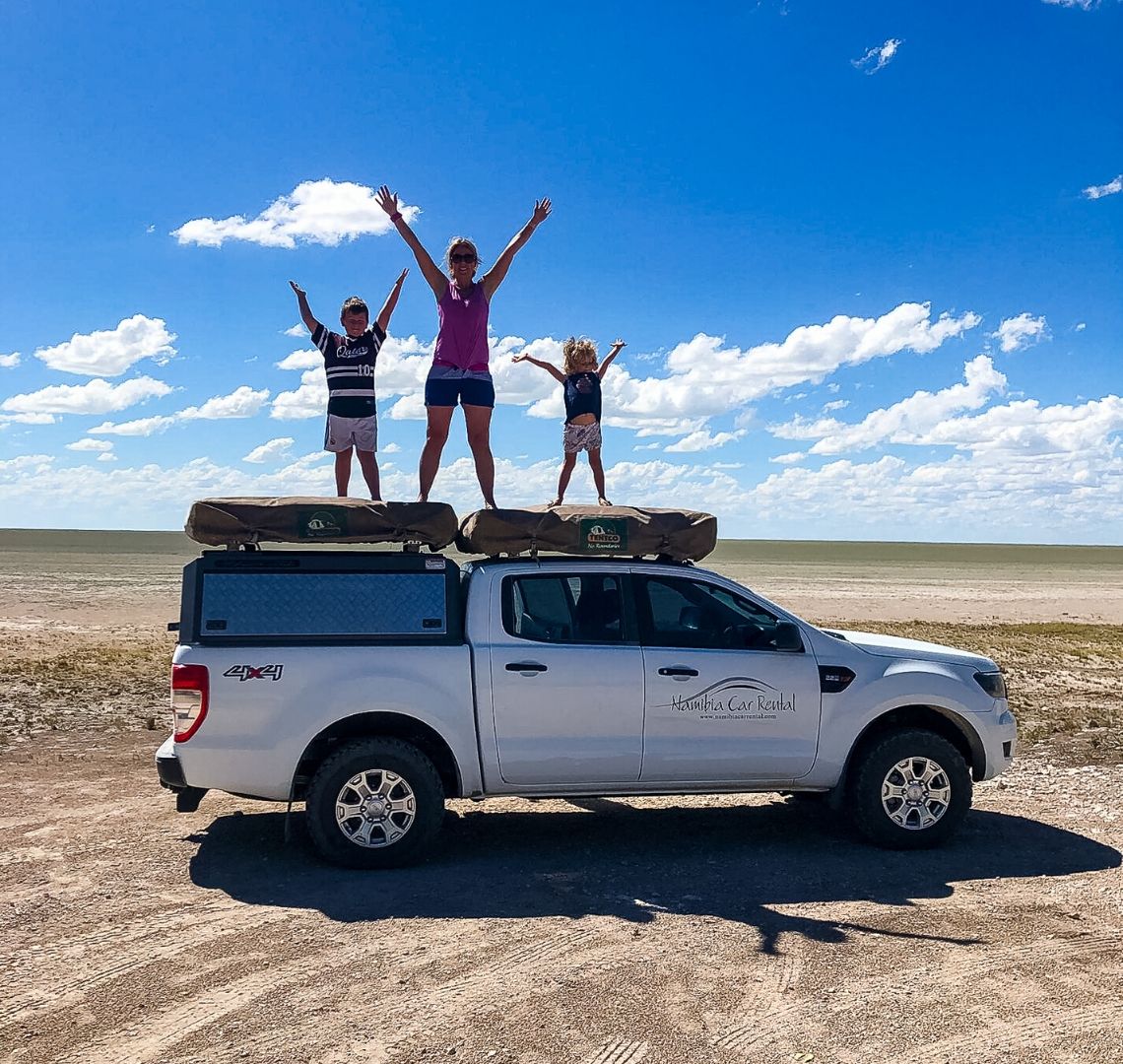 2 Week Namibia Itinerary - me and the 2 Things stood on top of our car with our arms in the air. Behind us is a rather grey and dusty looking salt pan. The sky is blue. 