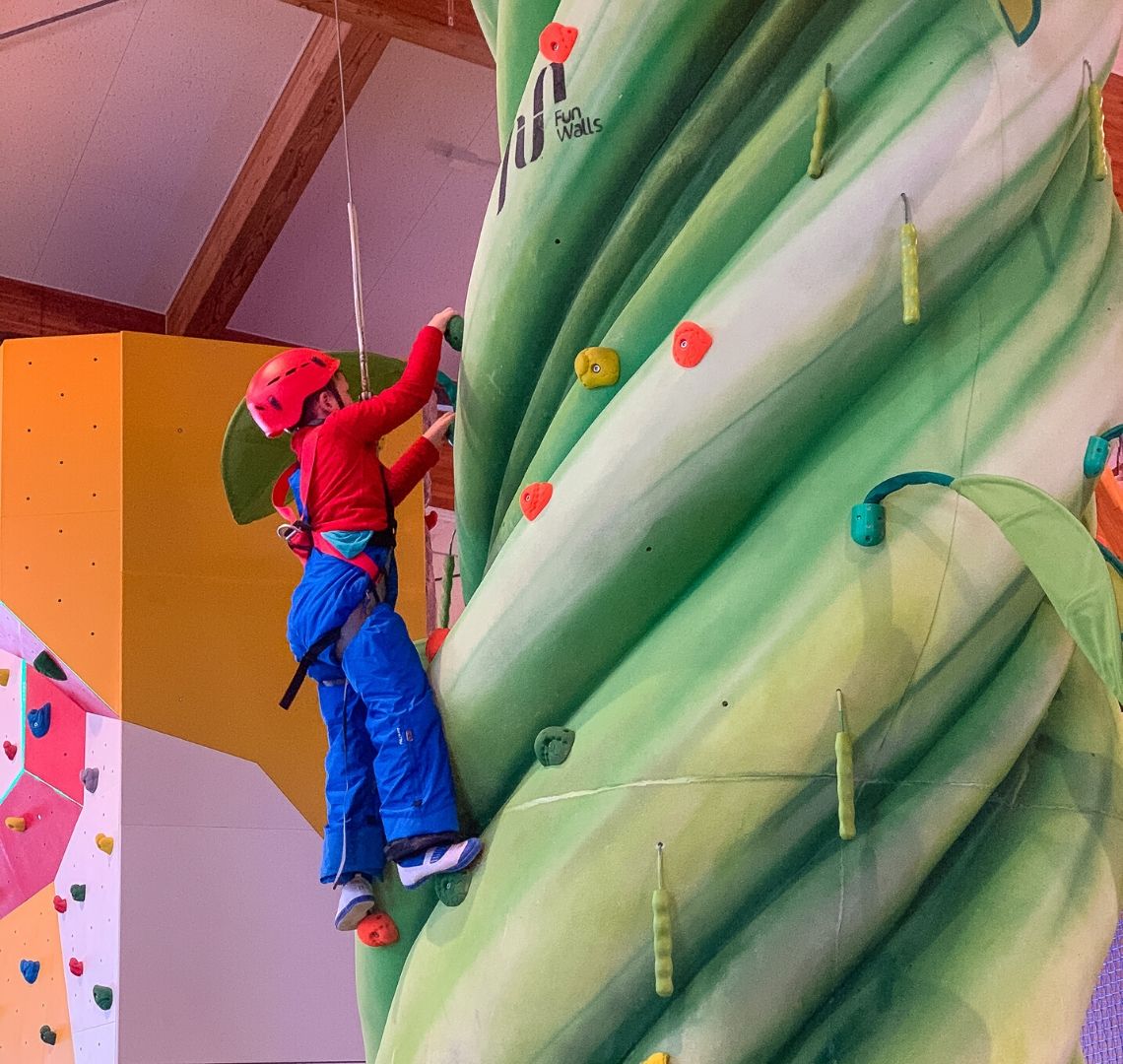 5 reasons to go family skiing in Niseko Thing 1 is wearing a bright red helmet, a bright red fleece and bright blue salopettes. He is near the top of a green climbing wall. 