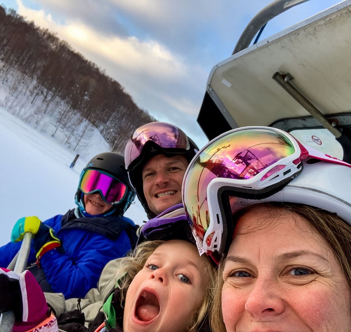 5 reasons to go family skiing in Niseko Wanderlust Family selfie on a ski lift - in order: me with my pink goggles wonky on my helmet, Thing 2 with pulling a funny face, Mr Wanderlust also with his goggles on his helmet and Thing 1 with rainbow goggles on, a black helmet and a bright blue jacket