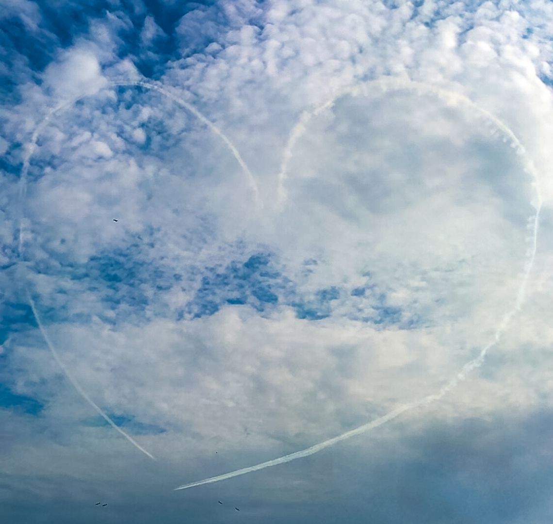 It Takes A Tribe - the sky is blue with lots of clouds. 2 red arrows planes have drawn a heart in white smoke trails