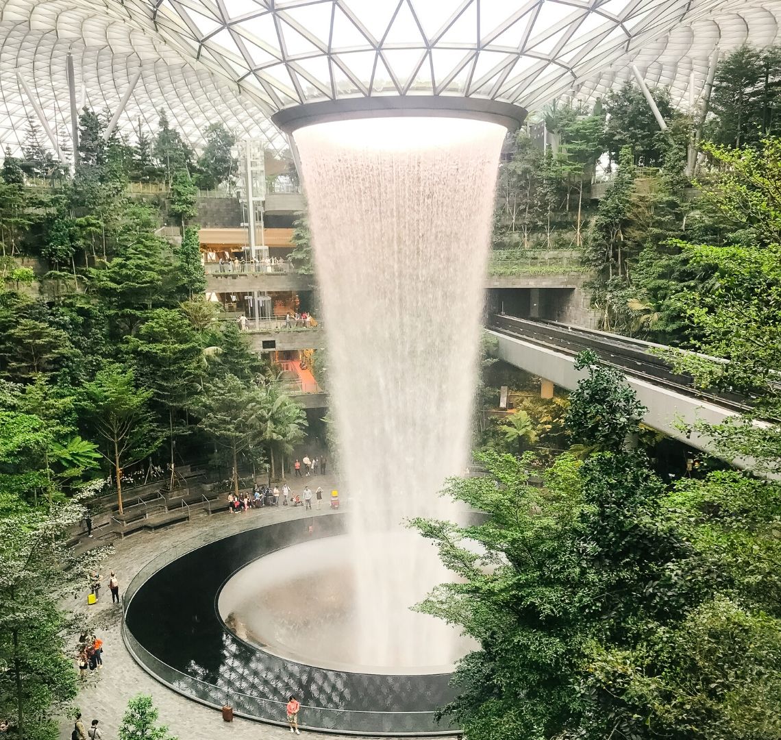 Jewel, Changi Airport, Singapore With Kids - the rain vortex thunders down in the Shiseido Forest Valley. There is a pathway all around where it disappears down to floors below and there are some people on it. There are trees to the left and right. On the right, the sky train track passes through. 