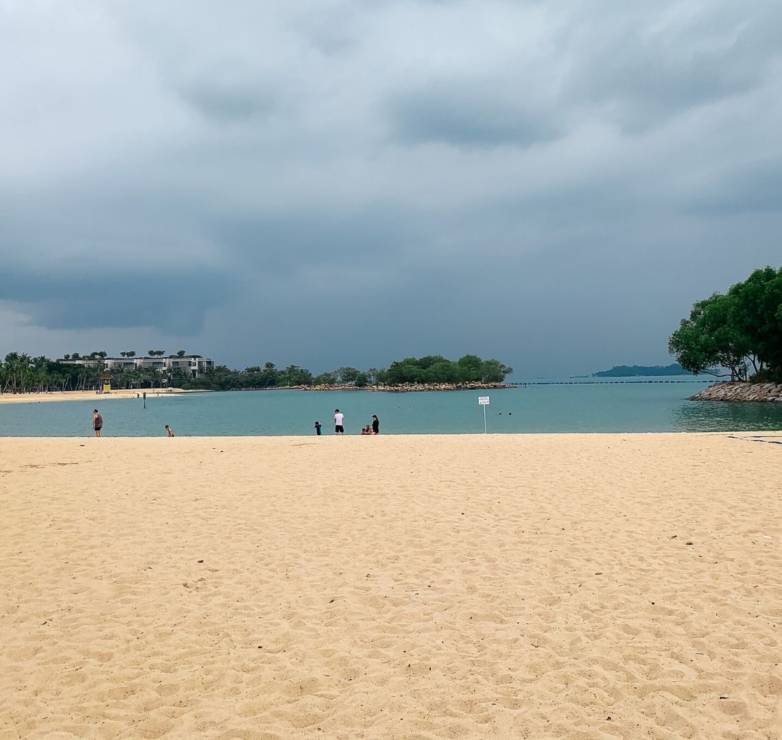 Things To Do In Sentosa With Kids - there is a long expanse of sand leading to a dark turquoise sea. There are a few people on the beach and a white sign is in the middle of the picture. To the right are some trees on a breakwater and to the left the beach bends out of sight and then curves round again where you can see some more trees and a building and another breakwater with trees on it. There is a grey island in the distance and the sky is stormy.