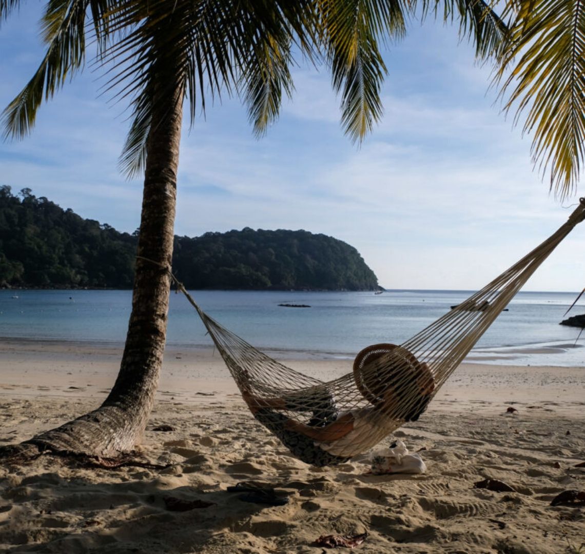 Best Secret Islands In Malaysia -  Pulau Tenggol - a lady in a hat sits in a hammock stung between 2 palm trees on a beach. The sea is completely calm and there is a dark green headland sticking out from the left.