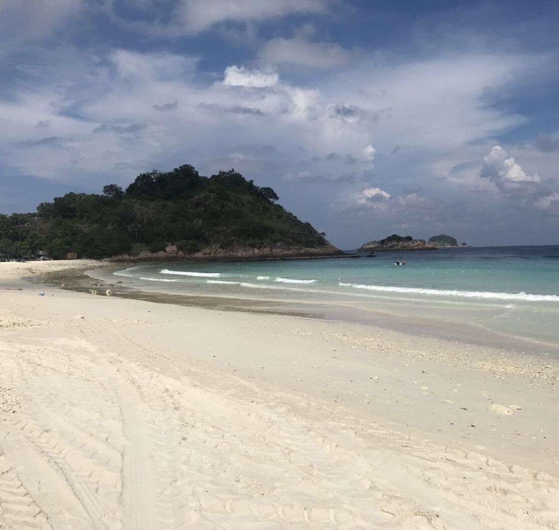 Best Secret Islands In Malaysia - more soft while sand curving round to to the left and then round to the right in the distance. At the end is a rocky outcrop with dark green trees and foliage. To the right and in the distance are a couple of tiny islands. The sky is blue at the top with some clouds but on the horizon is cloudier. 