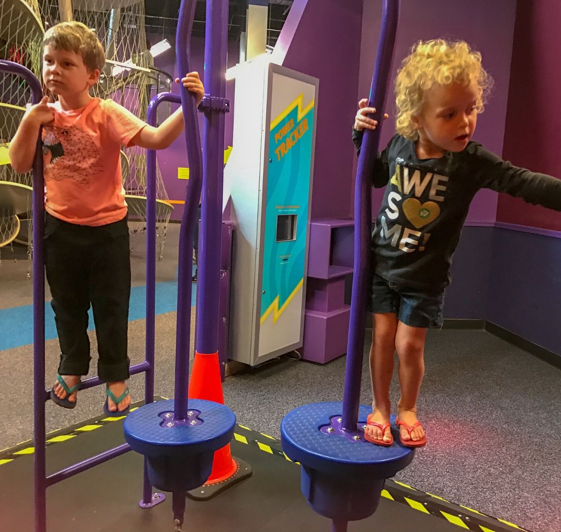 Thing 1 on the left in an orange t-shirt, black trousers and flip flops. Things 2 is in a black top with AWESOME written on it, denim shorts and flip flops. They are climbing and balancing on a purple exhibit at the Children's Museum of Houston