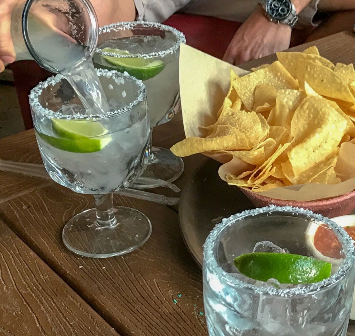Margaritas being poured in to salt rimmed glasses with ice and lime. There is a bowl of chips in the top right corner