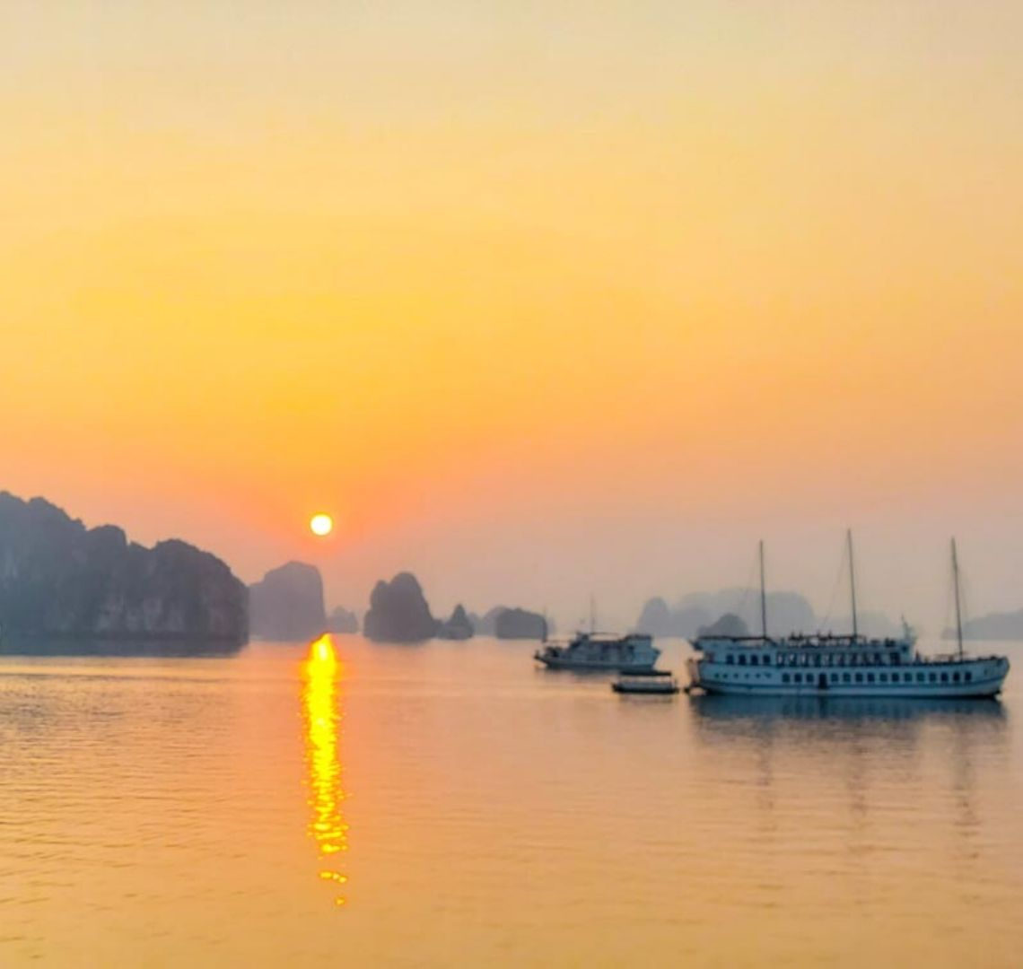 Best secret islands in Cambodia, Vietnam and Laos With Kids - the sun is setting in Bai Tu Long in Halong Bay. There are junk ships on the still water in the foreground and shadowy limestone rocks and islands in the background. The sky is bright orange and the tiny setting sun sets off an orange stripe in its reflection in the water.