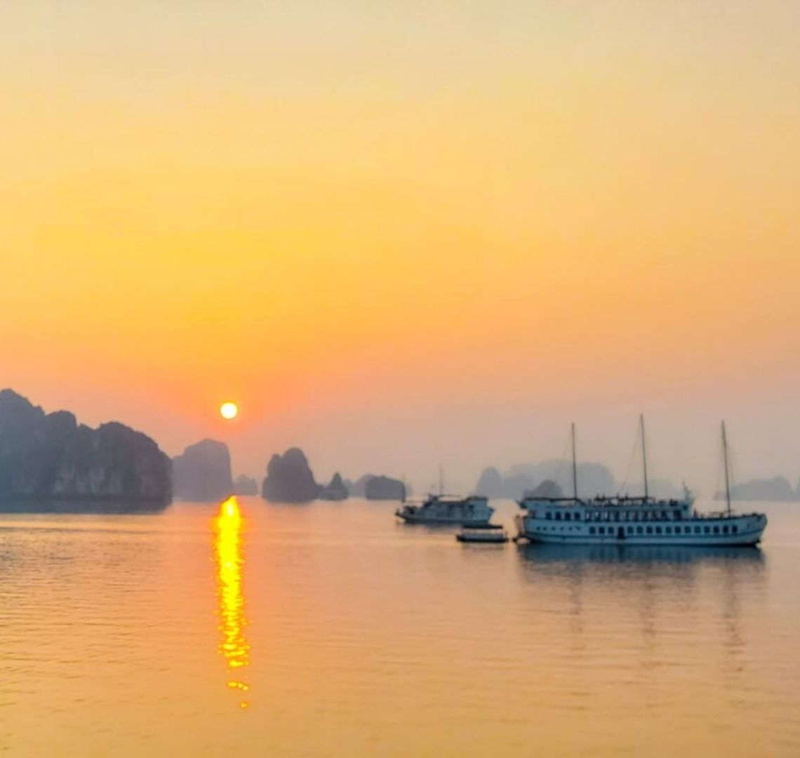 The sun is setting in Bai Tu Long in Halong Bay. There are junk ships on the still water in the foreground and shadowy limestone rocks and islands in the background. The sky is bright orange and the tiny setting sun sets off an orange stripe in its reflection in the water. 