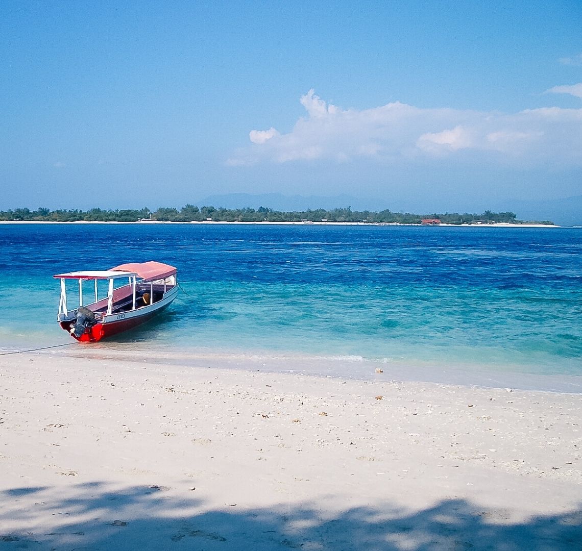 White white sand leads to turquoise water which then turns a deeper but bright blue. In the distance is another island with a thin white strip of sand and some trees and a building. In the shallows on this side, just pulled up to the beach is a red bottomed boat with white sides and a faded red roof. 