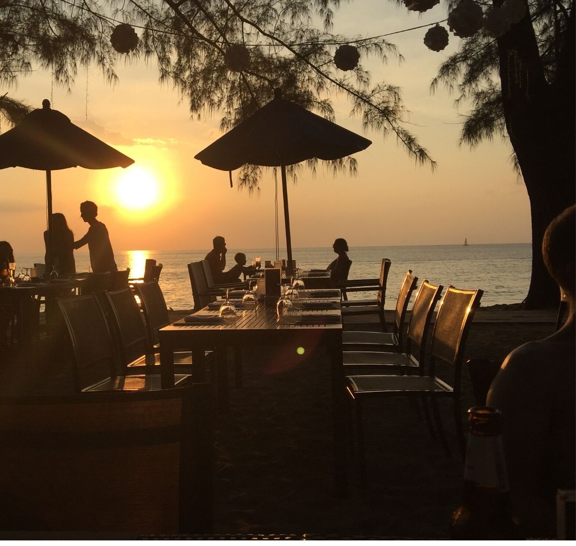 The best secret islands in Southeast Asia with kids - the sun is setting over the sea at the beach in Phuket, in teh foreground are shadowed tables with umbrellas with a few people dining, a tree and some hanging lights that aren't yet on.