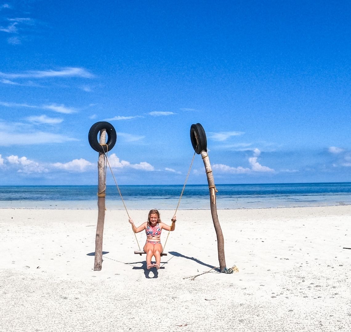 Siquijor - There is a vast expanse of white sand and beyond is deep blue sea and bright blue sky with just a few wispy clouds up high and some fluffier ones on the horizon, A little girl sits on.a swing in the centre of the photo - it is strung up between two large wooden poles with black tyres fixed to the top