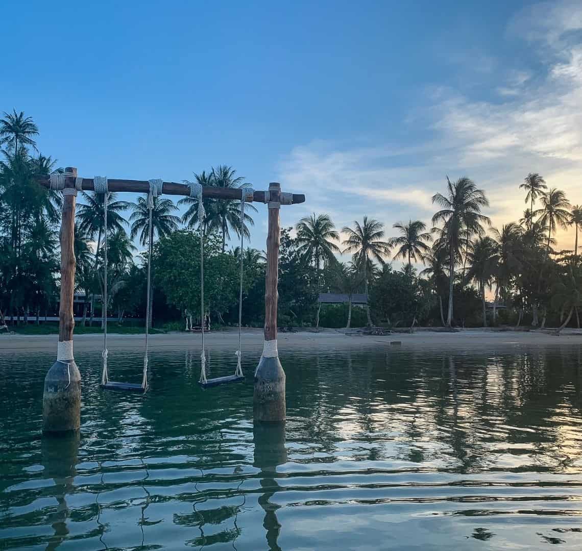 The Best Secret Islands In Southeast Asia - 2 swings in the ocean on the left with the beach in the background. The water is incredibly still and there are palm trees on the beach. The sky is blue with just a few wispy clouds on teh left. The moulds are illuminated by the last of a yellow sunset.