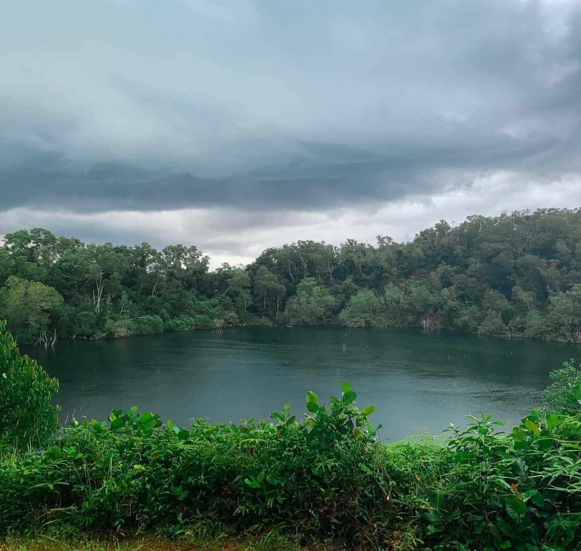 Bright green foliage at the bottom gives way to the dark water in teh quarry. It is surrounded by trees. The sky has dark storm clouds. 