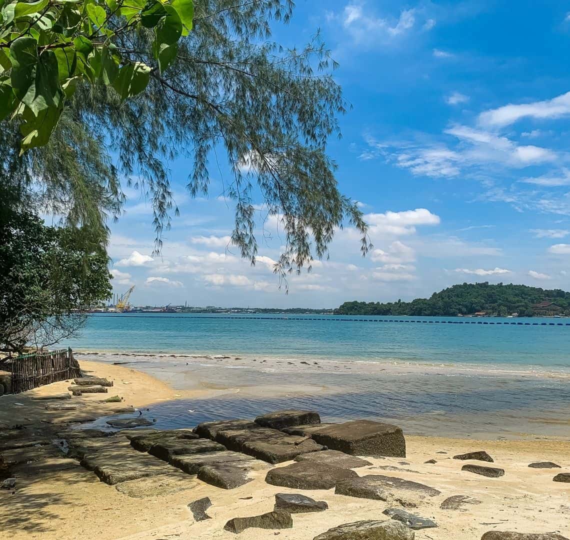 Coney Island Singapore - there is a yellow sandy beach at the bottom of the image with stepping stones on it. There are some tree branches hanging over from teh left. The turquoise sea is in the mid-ground and behind it the lush vegetation of Pulau Ubin. In the very far distance some cranes in Malaysia can be seen