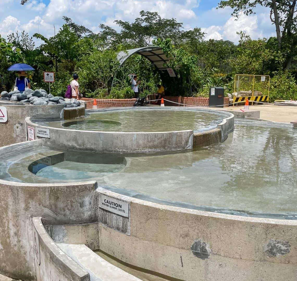 The image shows the cascading pools in Sembawang, They are circular and made of concrete. The water is clear. Behind the pools is a trellis that provides shade over some seats that are in front of a red brick wall and some tropical green plants. In the mid ground there are four people walking and sitting.