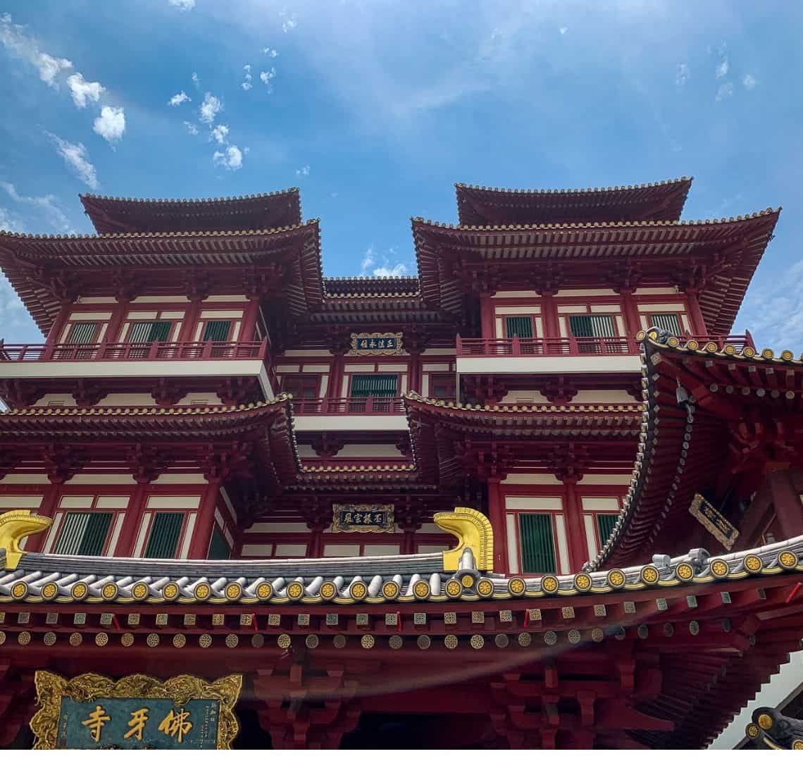 Buddha Tooth Relic Temple has Chinese roofs, white walls and lots of red edging with green shutters on the windows. above it the sky is blue with just a few clouds in the sky. 