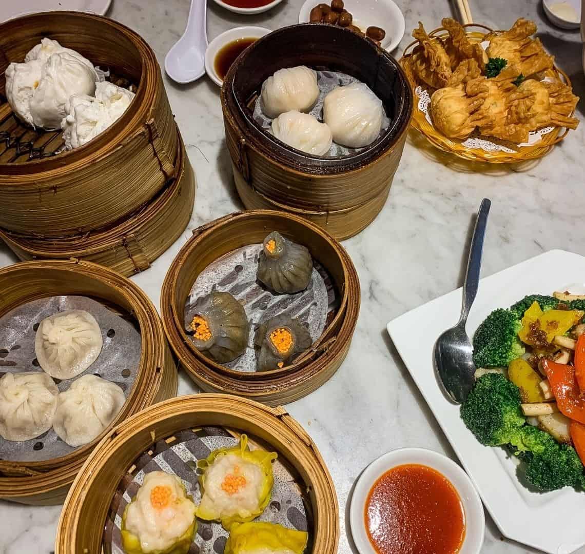 A selection of dumplings in bamboo steamers on a marble tabletop. On the bottom right is also a selection of colourful stir fried vegetables and at the top righter some fried wantons