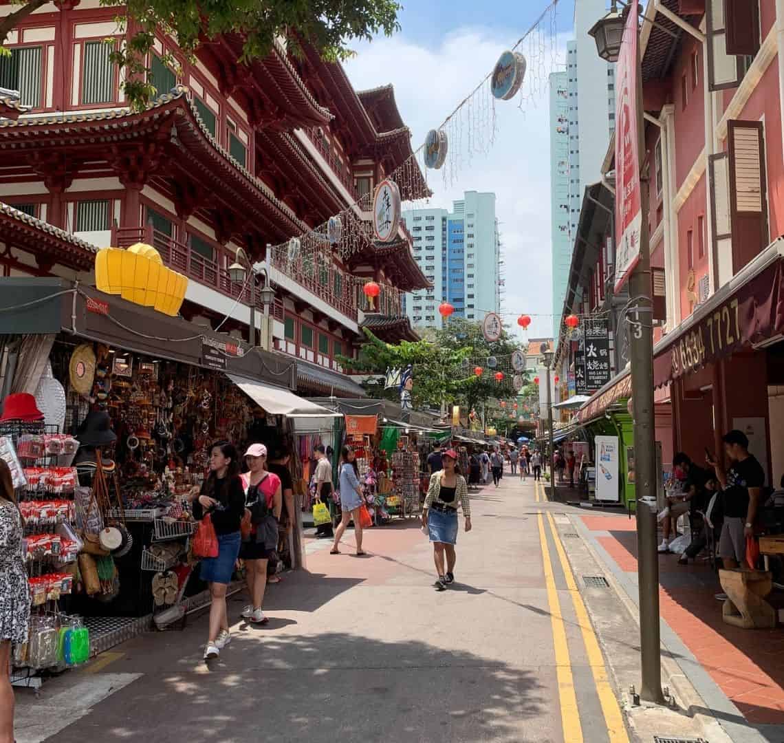 A Comprehensive Guide to Chinatown Singapore - Street view of some stalls along a street in Chinatown. They are selling all manner of tourist tat and there are a few tourists on the street. On the upper left above the stalls are the red walls of Buddha Tooth Relic Temple with its Chinese style roofs.