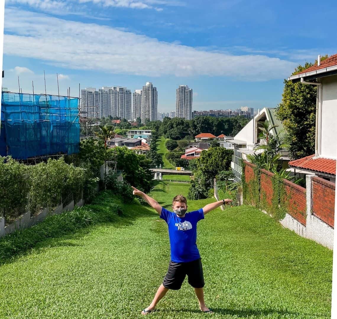 Think one in great shorts and a bright blue top stands at teh top of a green stretch of grass. On the left is green shrubbery and on teh rights some white walls with Spanish style terracotta grooves and shutters. There is also some scaffolding on the left. In the distance is a high rise, some smaller buildings and a blue sky