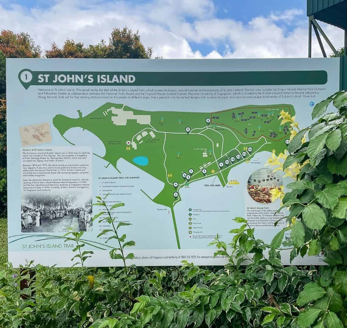 A map of St John's Island (photo of actual map sign) with some shrubbery and white clouds/blue sky behind it