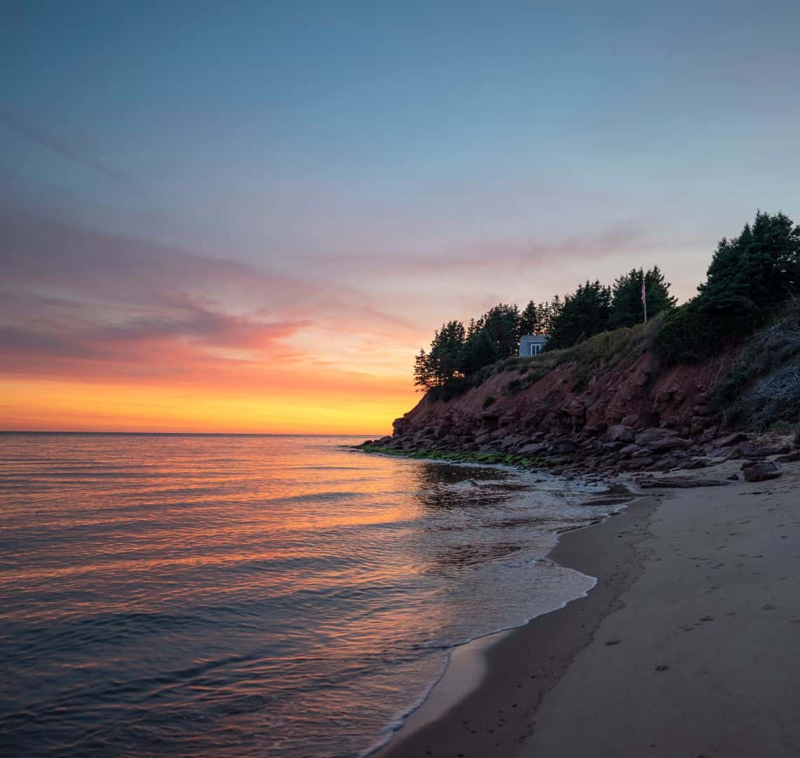 Sunset over the ocean with shallow waves on the left. On the bottom right is a sandy beach and behind it some low cliffs with some trees and a house on top