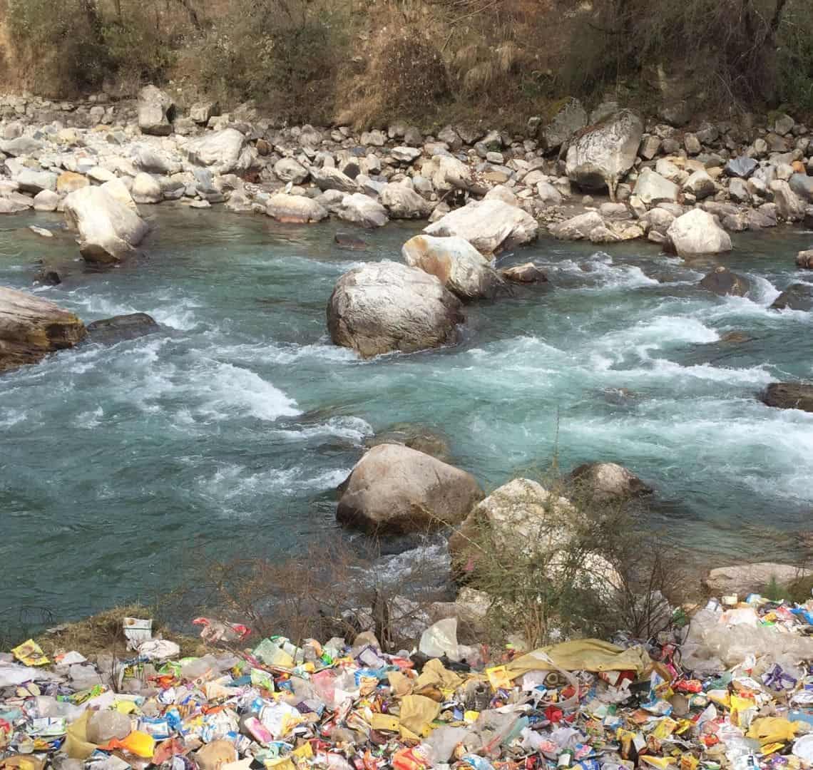 A river flowing over and around stones and boulders. The foreground is completely covered in trash. 