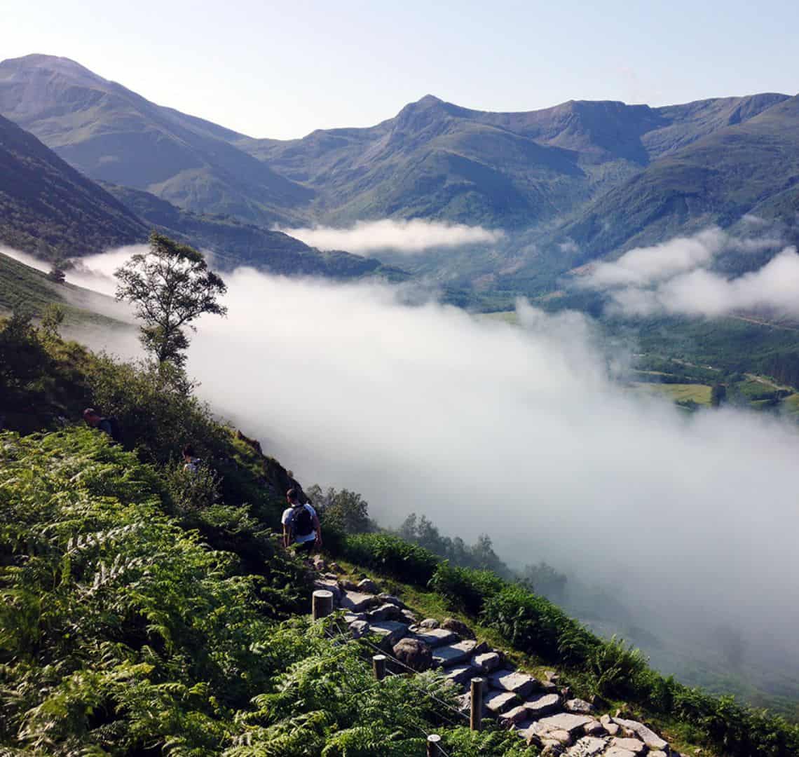 Lots of green hills with fog / mist in the valleys. There are three hikers in the foreground with day backpacks on.