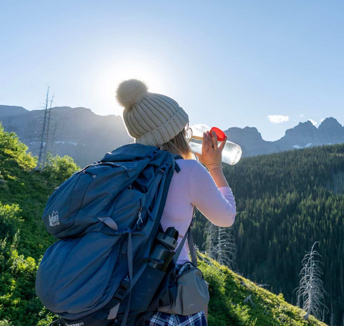 A woman in a cream bobble hat and light pink long sleeved top is drinking from a clear water bottle with a plastic lid. In the background is a forest and some mountains.
